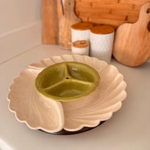May include: A three-sectioned serving dish with a light green center bowl and a cream-colored outer dish. The dish sits on a dark wooden base. Wooden cutting boards and white ceramic jars with wooden lids are in the background.