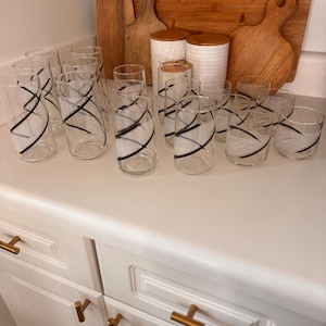 May include: A collection of clear drinking glasses with a black, abstract design. The glasses are arranged on a white countertop, alongside wooden cutting boards and white canisters with wooden lids. The glasses have a modern, minimalist aesthetic.