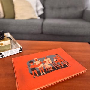 May include: An orange vintage children's book with an illustration of children in a classroom setting. The cover features a teacher and students, with a child painting at a desk. The book is on a wooden table.