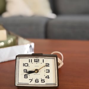 May include: A vintage Westclox dialite alarm clock with a black frame and a cream-colored face. The clock features black numerals, a gold-colored second hand, and the words "dialite" and "WESTCLOX" printed on the face. The clock is on a wooden surface.