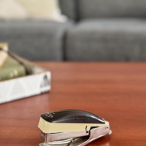 May include: A black and cream-colored stapler with silver metal components rests on a wooden surface. The stapler is a classic office supply, designed for fastening papers together. The word "ATES" is visible on the stapler.