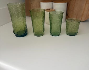 May include: A set of four green glass tumblers of varying heights, each featuring a textured, bark-like exterior pattern. The glasses are arranged in a row from tallest to shortest, showcasing a consistent translucent olive green color throughout.