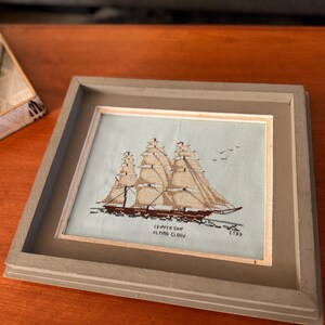 May include: Framed cross-stitch artwork of a clipper ship, featuring tan sails and a brown hull, set against a light blue background. The frame is a muted gray color. The text "CLIPPER SHIP FLYING CLOUD" is stitched below the ship.
