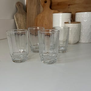 May include: Four clear glass drinking glasses with a ribbed design. The glasses are empty and sitting on a white surface.