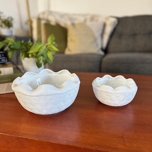 May include: Two white ceramic bowls with a diamond pattern and scalloped edges. The bowls are on a wooden table.