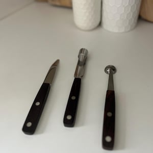 May include: Three kitchen tools with dark wooden handles and stainless steel blades. The tools include a paring knife, a melon baller, and a corer. Each tool has a silver rivet on the handle. The tools are arranged on a white surface.