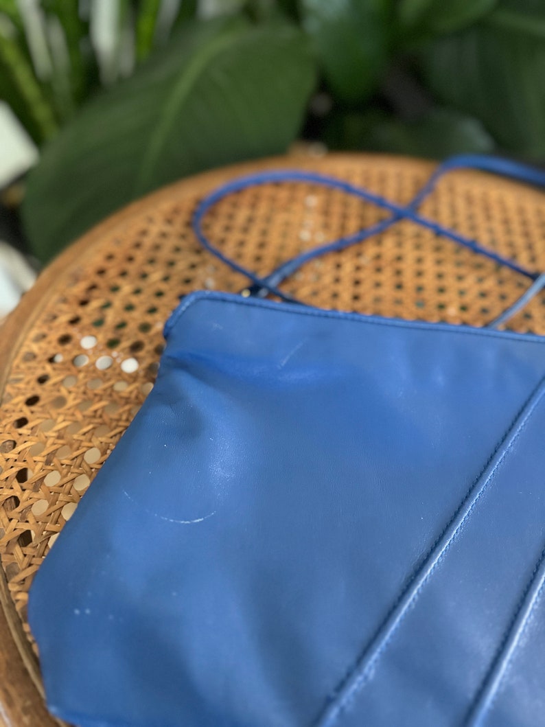 May include: A blue leather tote bag with a single strap. The bag has a smooth, slightly textured surface and is sitting on a woven chair.