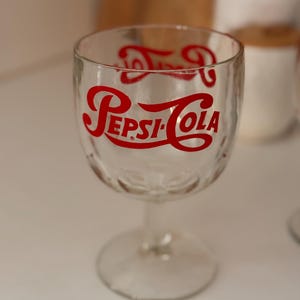 May include: Clear glass Pepsi-Cola goblet with a stem. The red Pepsi-Cola logo is printed on the front of the glass. The glass has a rounded bowl and a textured pattern near the top.