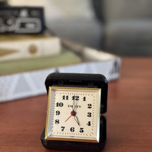 May include: A small, vintage travel alarm clock with a gold-colored square face and black numbers. The clock face reads "EQITY" and has red hands. The clock is housed in a black, hinged case.