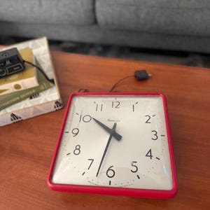 May include: A square, red-framed clock with a white face. Black hands point to the time, with black numerals marking the hours. The clock is labeled "SIMPLEX". The clock is on a wooden surface.