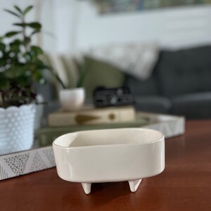 May include: White ceramic planter with four small legs. The planter is oval shaped and has a smooth, glossy finish.