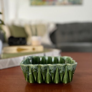 May include: A green ceramic planter with a scalloped edge and a textured, geometric design. The planter is made of glazed ceramic and has a light green glaze.