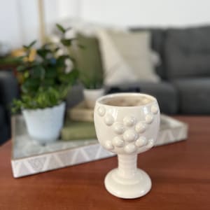 May include: White ceramic goblet with a textured surface. The goblet has a round base and a wide bowl with a raised pattern of small circles.