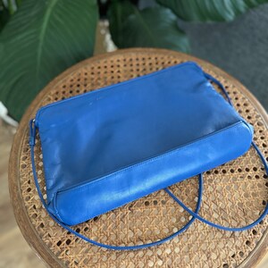 May include: A bright blue leather clutch bag with a long strap. The bag is sitting on a woven wicker surface.