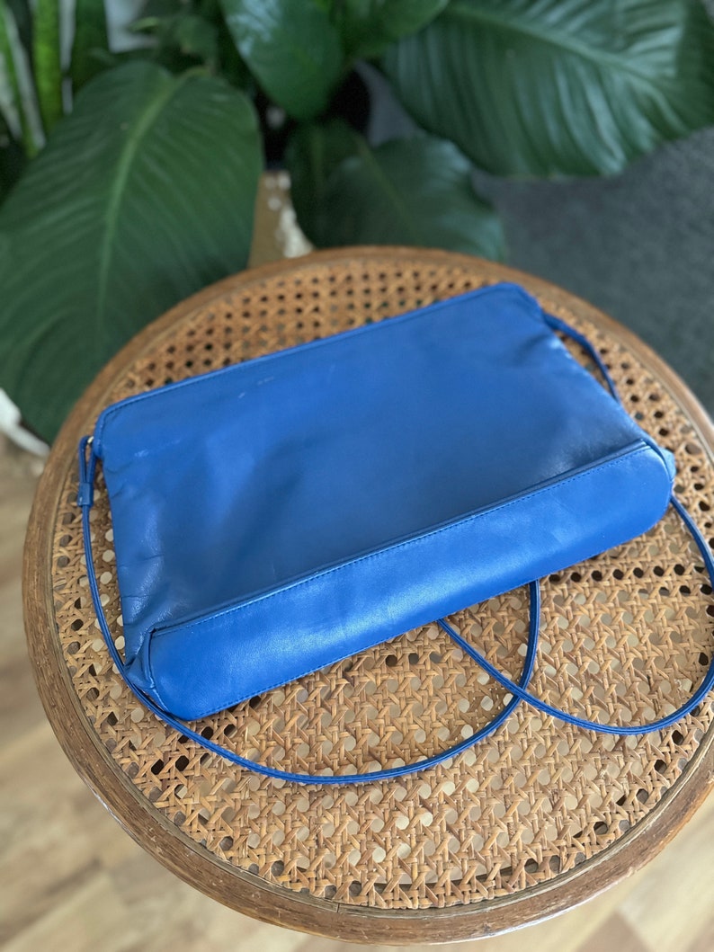 May include: A bright blue leather clutch bag with a long strap. The bag is sitting on a woven wicker surface.