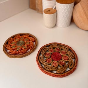 May include: Two round woven coasters with a floral design. The coasters are made of natural fibers in shades of brown, orange, and red. The coasters are on a white surface, with white ceramic containers and wooden cutting boards in the background.