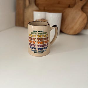 May include: A beige ceramic mug with a black handle and rim. The mug is printed with the words "KEY WEST" in a stacked, colorful design. The mug is sitting on a white surface.