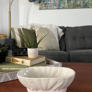 May include: A white ceramic bowl with a speckled finish. The bowl has a rounded oval shape with a slightly flared rim. The bowl is sitting on a wooden table.