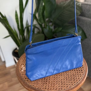 May include: A blue leather crossbody bag with a zipper closure and a long strap. The bag has a textured design with three lines running across the front.