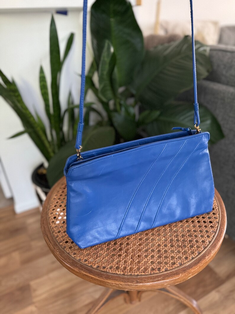 May include: A blue leather crossbody bag with a zipper closure and a long strap. The bag has a textured design with three lines running across the front.