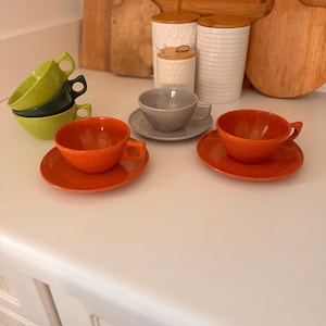 May include: A collection of colorful ceramic teacups and saucers. The set includes orange, gray, and green cups, with matching saucers. Wooden cutting boards and white canisters with wooden lids are in the background.