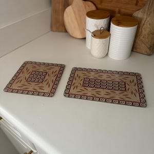 May include: Two rectangular beige and burgundy patterned placemats with a floral design. The placemats are on a white countertop, with wooden cutting boards and white ceramic containers with wooden lids in the background.