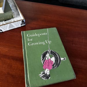 May include: A vintage book titled "Guideposts for Growing Up" with a green cover. The cover features an illustration of a person hanging upside down from a tire swing. The book is on a wooden surface.