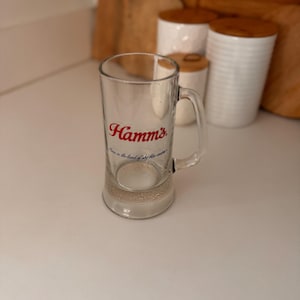May include: A clear glass beer mug with a handle. The mug has the word "Hamm's" in red script on the front. Below the logo is the phrase "Born in the land of sky blue waters" in blue script. The mug is on a white surface.