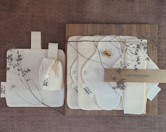 May include: A set of reusable cloth menstrual pads in a natural white colour, with a brown paper tag that says "princess&pea". The pads are arranged on a wooden surface with dried flowers.