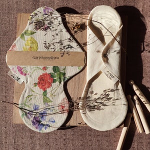 May include: Two organic cotton reusable cloth pads, one with a floral print and the other plain, folded and laid out on a wooden surface. The floral pad has the text "princess&pea organic cloth pads" printed on it.