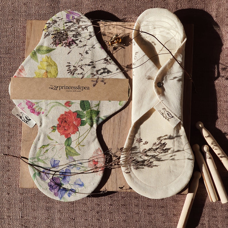 May include: Two organic cotton reusable cloth pads, one with a floral print and the other plain, folded and laid out on a wooden surface. The floral pad has the text "princess&pea organic cloth pads" printed on it.