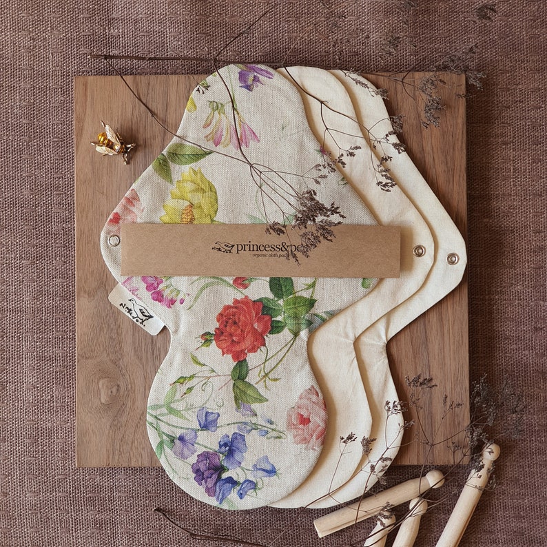 May include: Three reusable cloth pads with a floral print. The pads are made of organic cotton and are designed for menstrual hygiene. The pads are stacked on top of each other and have a brown label that says "princess&pea organic cloth pads".