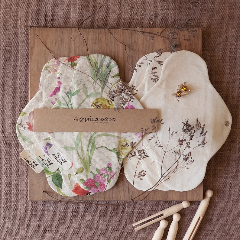 May include: A set of four reusable cloth pads with floral prints. The pads are made of organic cotton and are designed for menstrual hygiene. The pads are laid out on a wooden surface with a brown paper tag that reads "princess&pea organic cloth pads".