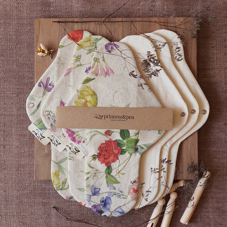 May include: A set of four reusable cloth menstrual pads with a floral print. The pads are made of organic cotton and are packaged in a brown paper envelope with the text "princess&pea organic cloth pads".