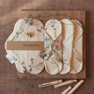 May include: Three organic cotton reusable cloth pads with floral print designs. The pads are folded and arranged on a wooden surface. The pads are labeled "princess&pea organic cloth pads".