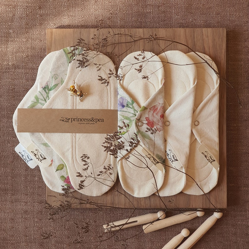 May include: Three organic cotton reusable cloth pads with floral print designs. The pads are folded and arranged on a wooden surface. The pads are labeled "princess&pea organic cloth pads".