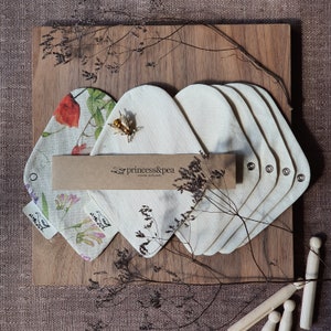 May include: A set of six organic cloth pads in a natural color, arranged on a wooden surface. The pads are made by princess&pea, a company that specializes in organic cloth pads. The pads are surrounded by dried flowers and a small bee charm.