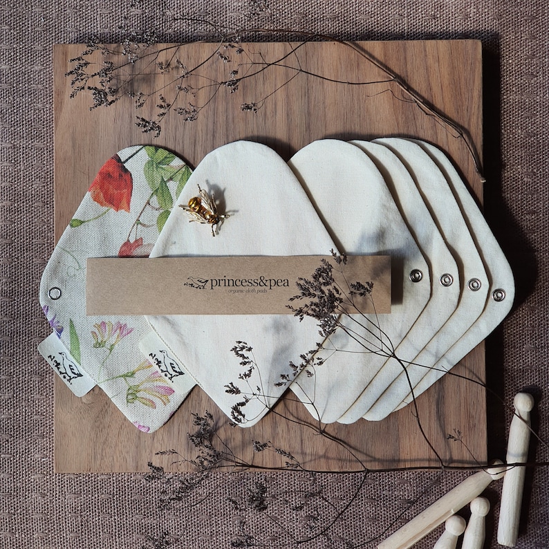 May include: A set of six organic cloth pads in a natural color, arranged on a wooden surface. The pads are made by princess&pea, a company that specializes in organic cloth pads. The pads are surrounded by dried flowers and a small bee charm.
