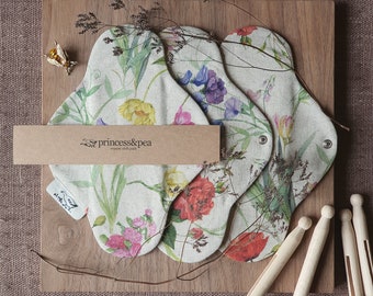 May include: Three reusable cloth pads with a floral print on a wooden surface. The pads are made by princess&pea, organic cloth pads.  There are also wooden clothespins on the surface.