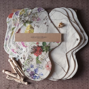 May include: Set of five reusable cloth menstrual pads with floral print. The pads are made of natural materials and are designed to be comfortable and absorbent. The pads are packaged in a brown box with the text "princess&pea" printed on it.