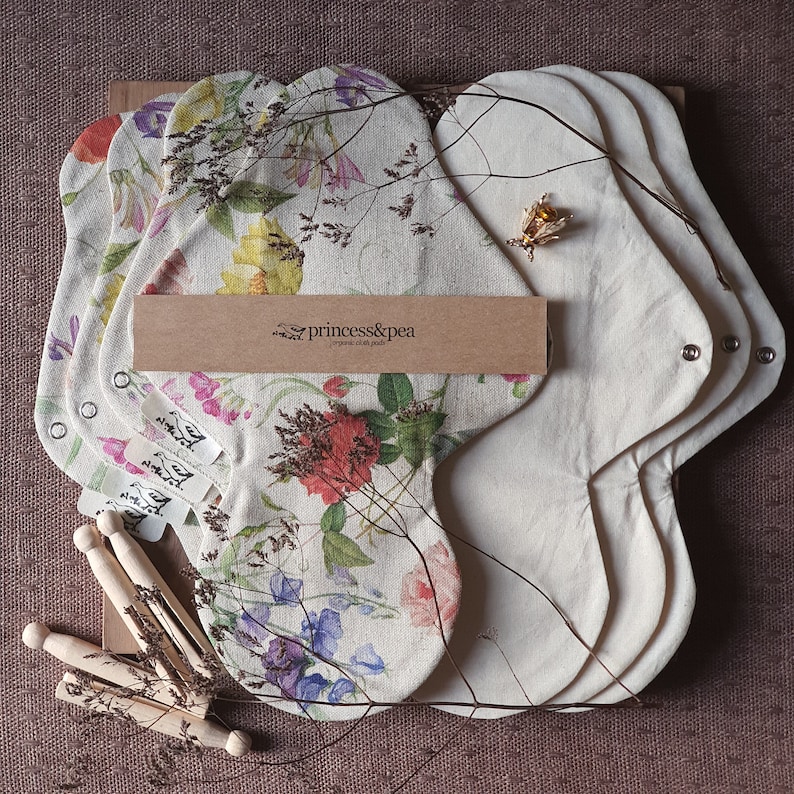 May include: Set of five reusable cloth menstrual pads with floral print. The pads are made of natural materials and are designed to be comfortable and absorbent. The pads are packaged in a brown box with the text "princess&pea" printed on it.
