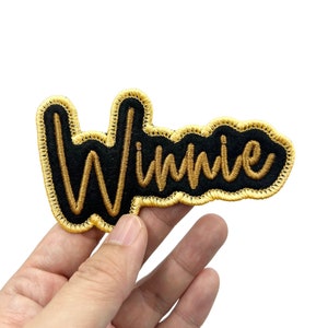 May include: A black felt patch with gold embroidered lettering that reads "Winnie".