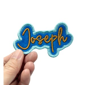 May include: A blue felt patch with a gold embroidered name "Joseph" in a cursive font.