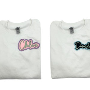 May include: Two white t-shirts with personalized names. The left shirt has a pink and green "Chloe" patch. The right shirt has a black and blue "Donald" patch.