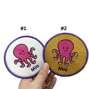 May include: Two round embroidered iron-on patches featuring a pink octopus design. One patch is white with a purple border and the other is gold glitter with a purple border. Both patches have the name "Mia" embroidered on them.