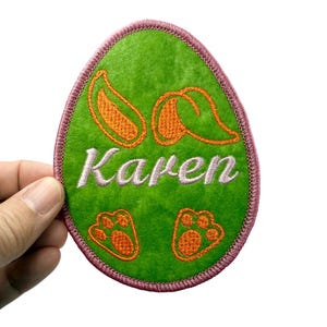Peut inclure: Patch en forme d'œuf de Pâques en feutre vert avec des coutures roses. Le patch est brodé avec des oreilles de lapin orange, des empreintes de pattes et le nom "Karen".