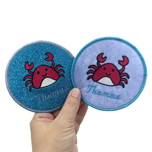 Peut inclure: Deux patchs ronds en tissu bleu et violet avec un motif de crabe rouge et le nom "Thomas" brodé sur chacun.