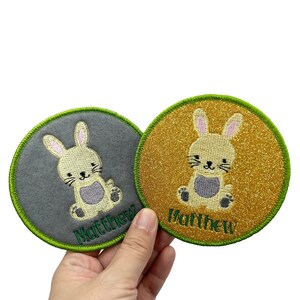 Peut inclure: Deux patchs thermocollants ronds avec une bordure verte. Chaque patch représente un lapin de dessin animé avec un corps brun clair, des pieds gris et des oreilles roses. Le lapin est assis. Les patchs sont sur un fond gris et or. Le nom "Matthew" est brodé en vert sur chaque patch.