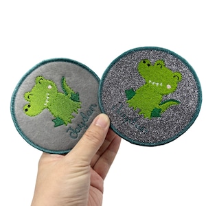 May include: Two round, personalized patches featuring a green cartoon alligator. One patch has a gray background with the name "Jaydan" embroidered in a cursive font. The other patch has a glittery gray background with the name "Cheyenne". Both patches have a teal border.
