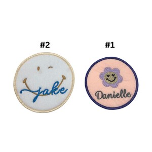 May include: Two embroidered iron-on patches. The first patch is pink with a purple flower with a smiley face and the name "Danielle" in grey embroidery. The second patch is white with a blue smiley face and the name "Jake" in gold embroidery.